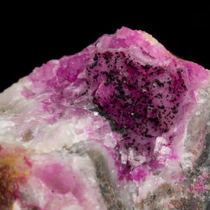 Puede incluir: Primer plano de un espécimen mineral con un vibrante tono rosa, salpicado de inclusiones negras. La roca tiene una estructura cristalina con áreas blancas y grises, sobre un fondo negro.