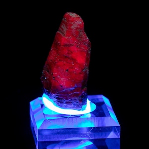 May include: A striking red gemstone, with a textured surface, is presented on a clear, square base, lit with blue and white light. The rough-cut stone is set against a black backdrop.