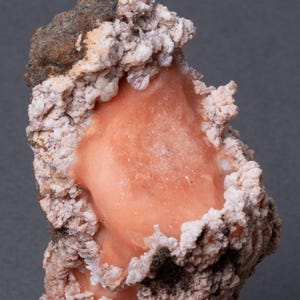 Peut inclure: Gros plan sur un spécimen minéral avec un cœur central de couleur pêche. Il est entouré d'une couche extérieure texturée, blanc cassé et marron. Le minéral a un aspect rugueux et cristallin.