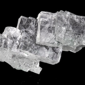 Puede incluir: Primer plano de una formación mineral cristalina transparente sobre un fondo negro. El mineral parece estar compuesto por bloques rectangulares apilados con una calidad ligeramente translúcida, mostrando estructuras internas.