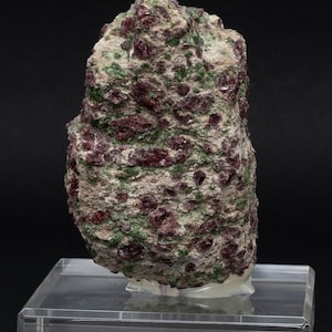 May include: A rough, irregularly shaped rock specimen, primarily gray with numerous deep red and green inclusions. The rock is displayed on a clear acrylic stand against a black background. The text on the stand reads "Pyrop. Chromdiopsid Ahem/NO".
