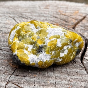 Pode incluir: Uma pedra natural arredondada com uma mistura de cores amarelo, branco e preto. A pedra tem uma superfície texturizada e está sobre uma superfície de madeira envelhecida. O padrão de cores único da pedra a torna um objeto decorativo.