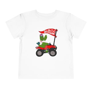 May include: White toddler t-shirt featuring a cartoon character riding a red ATV. The character, with green skin and a red hat, holds a banner that reads "Go Phils!" The ATV has black wheels and is set against a white background.