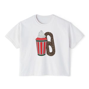 Op de afbeelding: Wit T-shirt met een grafisch ontwerp van een rood-wit gestreepte beker met slagroom en een bruine pretzel. De beker heeft een groene basis en de pretzel heeft witte spikkels. Een casual kledingstuk voor dagelijks gebruik.