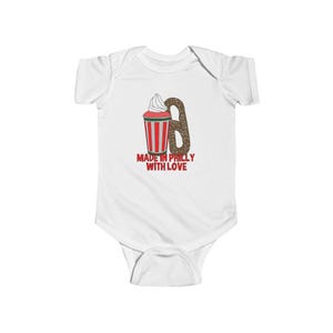 Op de afbeelding: Witte babybody met een afbeelding van een rood-wit gestreepte beker met slagroom, een pretzel en de tekst "MADE IN PHILLY WITH LOVE" in rood en groen.