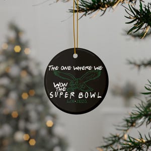 May include: Black round ceramic ornament with gold string, featuring the text "THE ONE WHERE WE WON THE SUPER BOWL LIX 2023" and a green eagle graphic. The ornament is hanging from a Christmas tree.