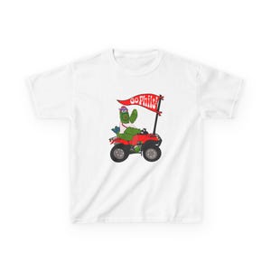 May include: White t-shirt featuring a cartoon character riding a red ATV. The character, wearing a hat and sunglasses, holds a flag that reads "GO PHILS!" The design is colorful and playful, perfect for fans of the Philadelphia Phillies.
