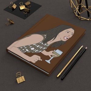 May include: Brown journal featuring an illustration of a woman holding a wine glass and pointing. The woman is wearing a patterned top. Gold and black pencils, and gold binder clips are also visible.