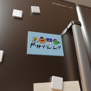 May include: A rectangular light blue magnet featuring four cartoon characters and the word "PHILLY" in black letters. The magnet is attached to a dark brown refrigerator door with white square magnets.