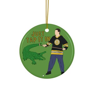 May include: Round green Christmas ornament with a cartoon illustration. The ornament features a person in a hockey jersey, a green alligator, and the text "JUST TAP IT IN". A gold string is attached for hanging.