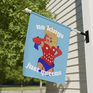 May include: A light blue flag with the text "no kings just queens" and a cartoon illustration of a drag queen in a red corset with white stars, red gloves, and blue shorts. The flag is hanging outside.
