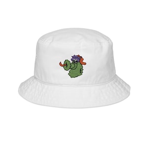 May include: White bucket hat with an embroidered green monster design wearing a purple hat and orange visor. The hat has a wide brim and a rounded crown. The monster has a red tongue sticking out.