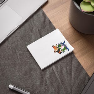 May include: A white rectangular notepad featuring a colourful graphic of four cartoon characters standing together. The characters include an orange figure in a jersey, a white and black figure, a green figure, and a blue figure in a dark outfit.