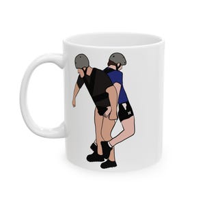 Könnte beinhalten: Weiße Keramik-Tasse mit einer Cartoon-Illustration von zwei Figuren in schwarzer und blauer Sportkleidung, die Helme tragen. Die Figuren befinden sich in einer Ringkampfposition. Die Tasse hat einen weißen Griff und Innenraum.