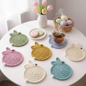 May include: A collection of handmade, crocheted bunny-shaped coasters in various pastel colors, including green, pink, yellow, blue, and cream. Each coaster features bunny ears and a small white pom-pom tail. The coasters are arranged on a white table.