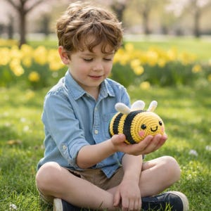 May include: A crocheted bee toy with yellow, black, and white details. The bee has a smiling face and small wings. The toy is held in a person's hands, with a blurred background of green grass and yellow flowers.