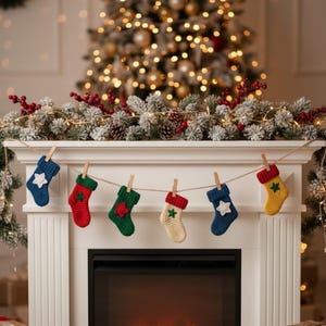May include: A garland of six miniature knitted Christmas stockings in various colors, including blue, red, green, and yellow, each adorned with a star. The stockings are suspended by clothespins on a string above a fireplace, with a decorated Christmas tree in the background.