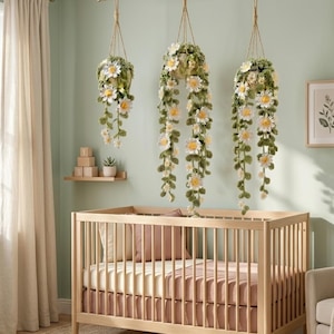 May include: Three hanging crochet flower baskets with long, trailing vines of white and yellow flowers and green leaves. The baskets are suspended from the ceiling above a wooden crib with a pink and cream bedding set. The room has a soft, neutral color palette.