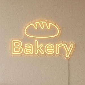 May include: A neon sign with the word "Bakery" in a yellow glow, with a bread loaf icon above the text. The sign is mounted on a cream-colored wall. A glass display case with baked goods is in the foreground.