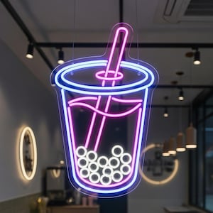 May include: A neon sign in the shape of a bubble tea cup, with a blue and pink outline. The cup contains white spheres, representing the tapioca pearls. The sign is suspended from the ceiling, with a black track lighting system visible.