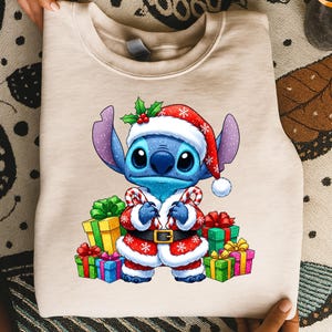 Puede incluir: Sudadera beige con un personaje de dibujos animados, Stitch, vestido con un traje de Pap&aacute; Noel. El personaje azul lleva un gorro y abrigo de Pap&aacute; Noel rojo y blanco, sosteniendo bastones de caramelo. Regalos coloridos rodean al personaje, tema navide&ntilde;o.