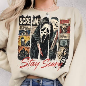 Puede incluir: Una sudadera beige de cuello redondo con un estampado gráfico del personaje de la película Scream con una túnica negra con capucha sosteniendo cuchillos, rodeado de paneles estilo cómic con texto que incluye Scream, Horror Tales y Stay Scary en rojo.