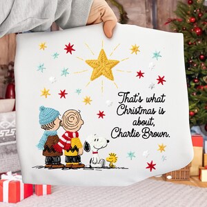 Może przedstawiać: Biała poszewka na poduszkę o tematyce świątecznej z haftowanymi wizerunkami Charliego Browna, Snoopy'ego i Woodstocka. Poszewka ma dużą złotą gwiazdę i napis "That's what Christmas is about, Charlie Brown."