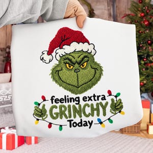 May include: White pillowcase with an embroidered Grinch design wearing a Santa hat. The Grinch is green with yellow eyes and a grumpy expression. Text reads "feeling extra GRINCHY Today" in green and white.