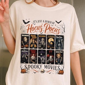 May include: A cream-colored t-shirt featuring a graphic print with the text IT'S JUST A BUNCH OF HOCUS POCUS and SPOOKY MOVIES. The design includes illustrations of ten classic horror movie VHS covers, spiderwebs, bats, and two orange pumpkins.