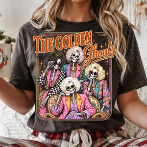 A woman wearing a black t-shirt with a graphic design featuring a group of skeletons dressed in colorful outfits, with the text "The Golden Ghouls" above them.