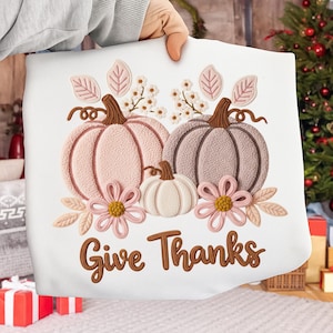 May include: White pillow with an embroidered design featuring three pumpkins in shades of pink and taupe, with floral accents and the words "Give Thanks" in brown script. A festive autumn-themed decorative item.