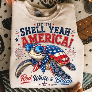 Könnte beinhalten: Ein beigefarbenes Sweatshirt mit Rundhalsausschnitt und einer Grafik einer Meeresschildkröte mit rot-weiß-blauer Sonnenbrille und passendem Halstuch. Der Panzer hat ein Flaggenmuster. Text: EST. 1776, SHELL YEAH AMERICA! und Red, White & Beach.