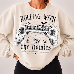 Puede incluir: Una sudadera de cuello redondo color crema con un estampado gráfico negro de dos manos liando un cigarrillo con personajes de películas de terror. El texto dice ROLLING WITH the homies GOOD FRIENDS GOOD TIMES BAD DECISIONS con murciélagos.