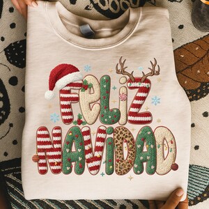 K&ouml;nnte beinhalten: Beiges Sweatshirt mit dem Schriftzug "Feliz Navidad" in festlichem Design. Der Text zeigt einen Weihnachtsmannhut, Rentiergeweihe und eine Mischung aus roten, gr&uuml;nen und Leopardenmustern, perfekt f&uuml;r die Weihnachtszeit.