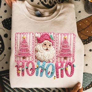 May include: A beige sweatshirt featuring a festive Christmas design. The design includes pink Christmas trees, a Santa Claus, and the text "HOHOHO" in a playful font. The design is embroidered with a textured appearance.