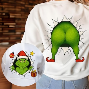 May include: White sweatshirt featuring a green cartoon character design. The back of the sweatshirt shows the character's green bottom and legs with red shoes bursting through a wall. The front shows the character's face wearing a Santa hat.