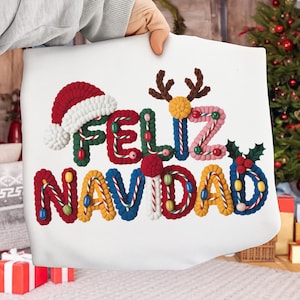 Puede incluir: Un cojín blanco con las palabras "FELIZ NAVIDAD" en letras coloridas y texturizadas. El diseño incluye un gorro de Papá Noel, cuernos de reno y acebo. Las letras son rojas, verdes, azules, amarillas y rosas.