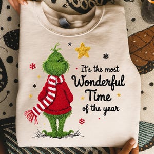 May include: A beige sweatshirt featuring a green Grinch character wearing a red scarf and sweater. The text "It's the most Wonderful Time of the Year" is printed on the front, along with star and snowflake graphics. A festive holiday design.