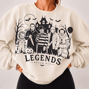 Puede incluir: Una sudadera de cuello redondo color crema con un estampado gráfico negro de personajes icónicos de películas de terror, una casa encantada, murciélagos y una calabaza. El texto "LEGENDS NEVER DIE" aparece debajo, junto a una lápida con "R.I.P.".
