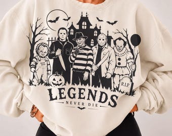 May include: A cream-coloured crewneck sweatshirt featuring a black graphic print of iconic horror movie characters, a haunted house, bats, and a pumpkin. The text "LEGENDS NEVER DIE" is printed below the characters, with a tombstone marked "R.I.P."