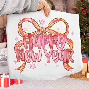 May include: A white pillow with a pink and gold bow design and the words "Happy New Year" in pink. The design includes pink snowflakes. The pillow is held by a person.