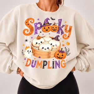 Może przedstawiać: Kremowa bluza z okrągłym dekoltem z nadrukiem graficznym z napisem "Spooky DUMPLING". Wzór przedstawia trzy uśmiechnięte pierożki w bambusowym koszyku do gotowania na parze, otoczone dyniami, duszkiem i małymi gwiazdkami.