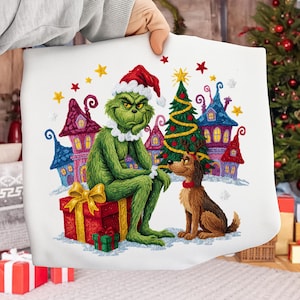 May include: A white Christmas cushion featuring a green Grinch character in a Santa hat, seated on a red gift box. A brown dog sits beside him. Colourful houses and a Christmas tree are in the background. Yellow and red stars are scattered around the image.