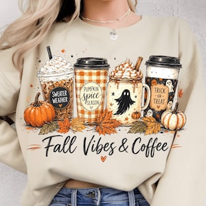 Puede incluir: Una sudadera beige con un gráfico de cuatro tazas de café, calabazas y hojas de otoño. Las tazas tienen las etiquetas Sweater Weather, Pumpkin Spice Season y Trick-or-Treat. El texto Fall Vibes & Coffee está impreso en negro debajo.