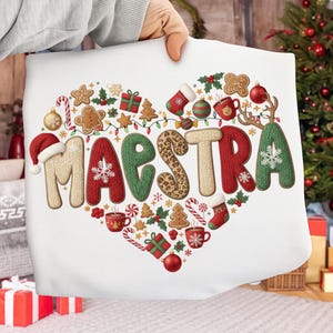 May include: A white pillow with a heart-shaped design filled with Christmas-themed illustrations and the word "MAESTRA" in festive lettering. The design includes gingerbread men, ornaments, and candy canes.