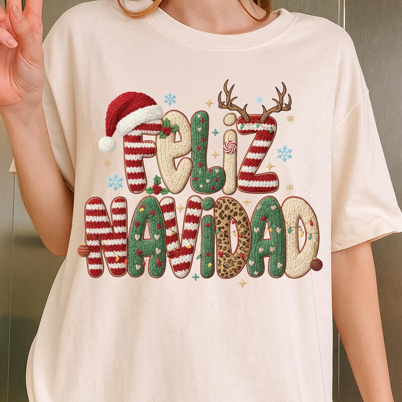 K&ouml;nnte beinhalten: Cremefarbenes T-Shirt mit dem spanischen Ausdruck "Feliz Navidad". Das festliche Design umfasst einen Weihnachtsmannhut, Rentiergeweih und dekorative Elemente in Rot, Gr&uuml;n und Leopardenmuster.