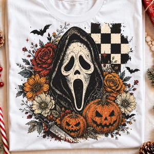May include: A white t-shirt featuring a graphic print of a ghost-like mask in a dark hood, surrounded by red and orange roses, white flowers, two jack-o'-lanterns, a knife, bats, and a black and white checkered pattern in the background.