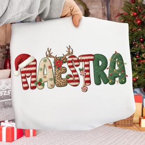May include: A white pillow with the word "MAESTRA" embroidered in festive Christmas-themed designs. The letters are decorated with a Santa hat, reindeer antlers, leopard print, holly, and candy canes. The letter "R" is adorned with Christmas lights.