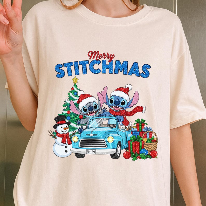 Puede incluir: Camiseta color crema con un gr&aacute;fico festivo. El dise&ntilde;o incluye dos personajes azules con aspecto de extraterrestres con gorros de Pap&aacute; Noel, una camioneta azul, un &aacute;rbol de Navidad y un mu&ntilde;eco de nieve. El texto "Merry Stitchmas" est&aacute; arriba.