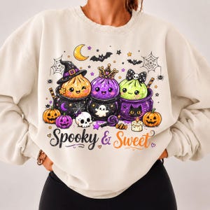 May include: A cream-colored sweatshirt featuring a graphic print of three colorful, smiling creatures in cauldrons, surrounded by bats, spiderwebs, a crescent moon, pumpkins, a skull, and a candle, with the text Spooky & Sweet printed below.